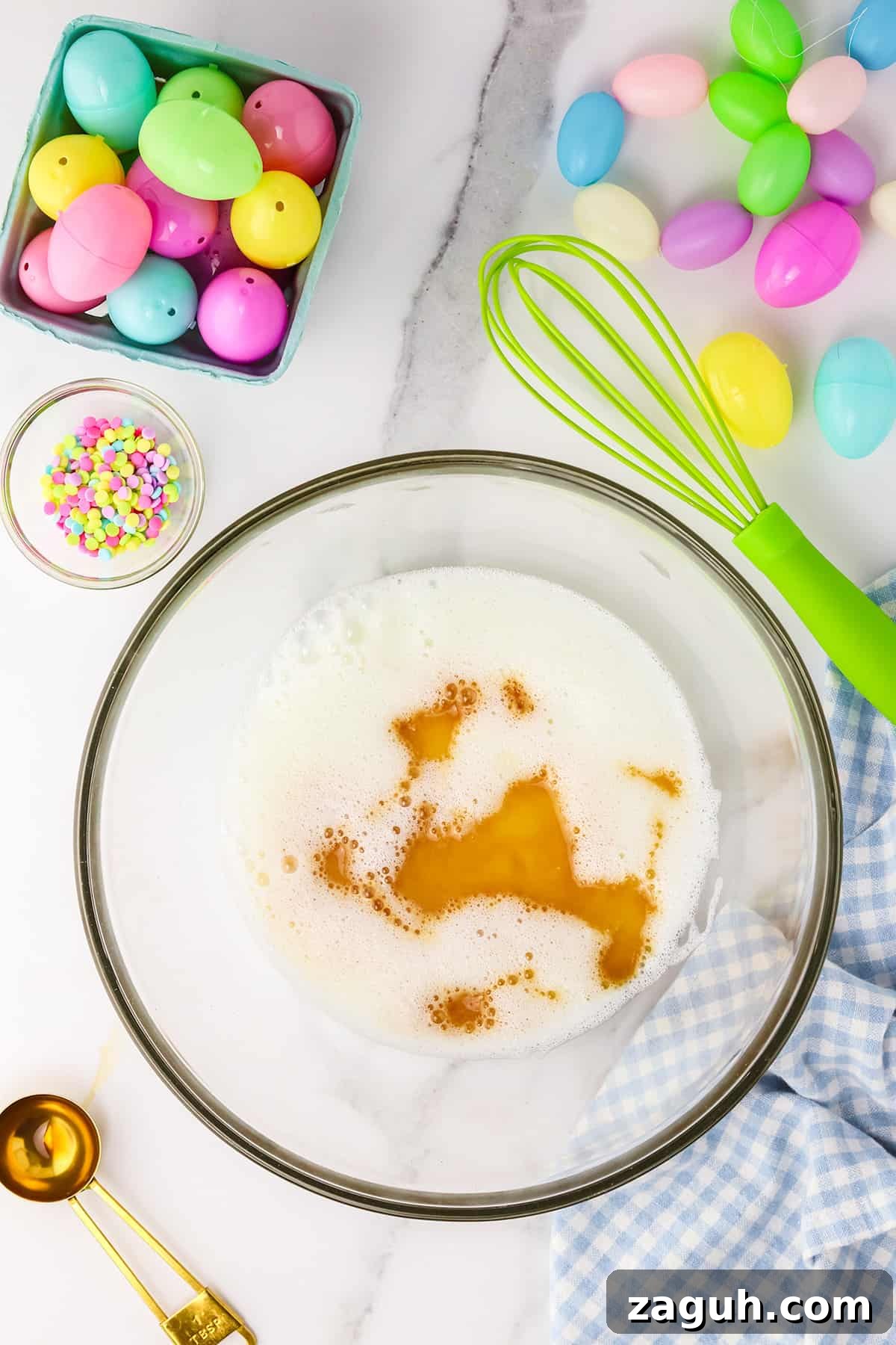 How to Make Copycat Cadbury Creme Egg Candy at Home 7 Baking preparation with colorful Easter Copycat Creme Egg candy, whisk, and ingredients on a marble countertop.