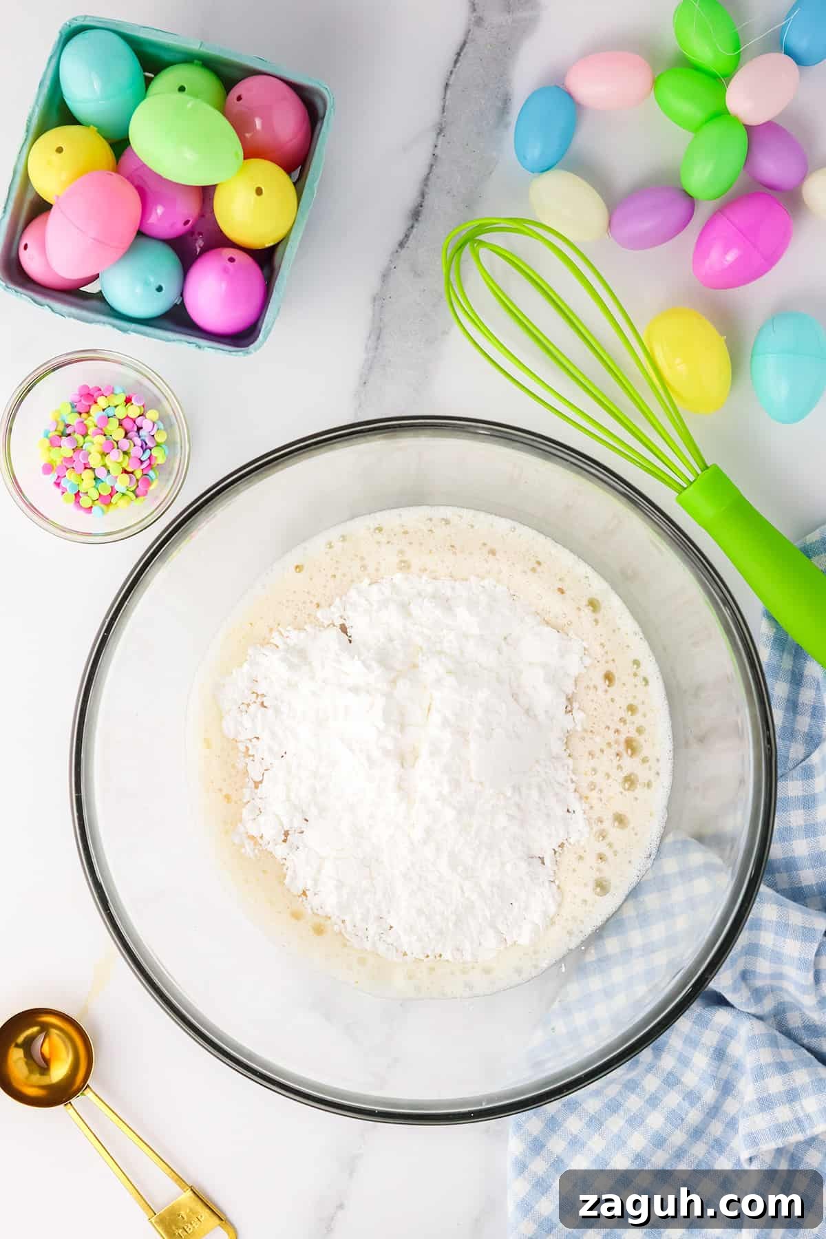 How to Make Copycat Cadbury Creme Egg Candy at Home 8 Baking ingredients in a bowl with colorful Easter Candy and a whisk on a marble countertop.