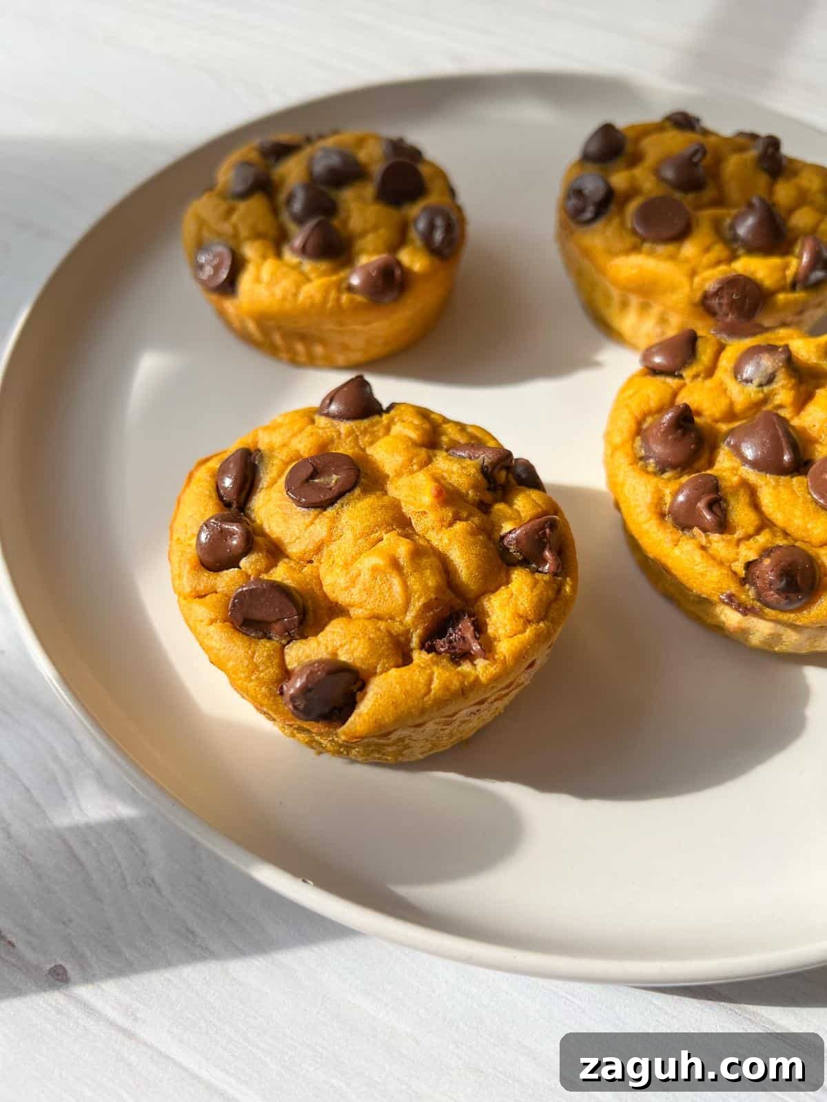 Moist Pumpkin Spice Muffins with Simple Ingredients 2 Four pumpkin-flavored muffins with chocolate chips on a round, beige plate. The muffins are arranged closely together, with sunlight casting soft shadows on the white surface beneath the plate.