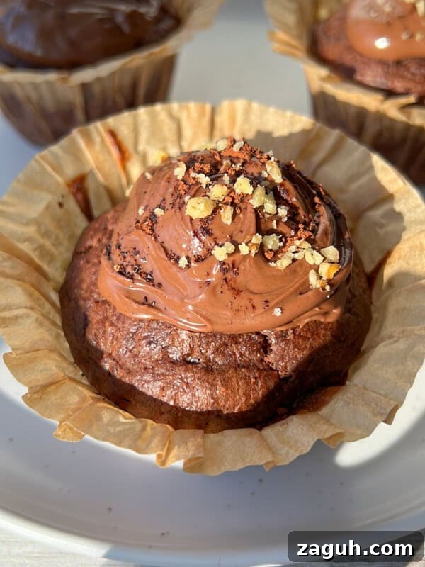Moist Pumpkin Spice Muffins with Simple Ingredients 12 Close-up of a chocolate cupcake in a paper liner, topped with chocolate frosting and sprinkled with chopped nuts. Two more decorated cupcakes are visible in the background on a white plate.