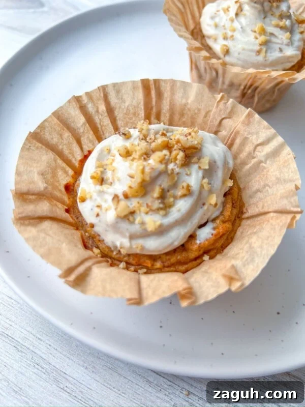 Moist Pumpkin Spice Muffins with Simple Ingredients 13 A Healthy Carrot Cake Muffin with white frosting and chopped nuts sits in a brown paper liner on a white plate, with another similar muffin blurred in the background.