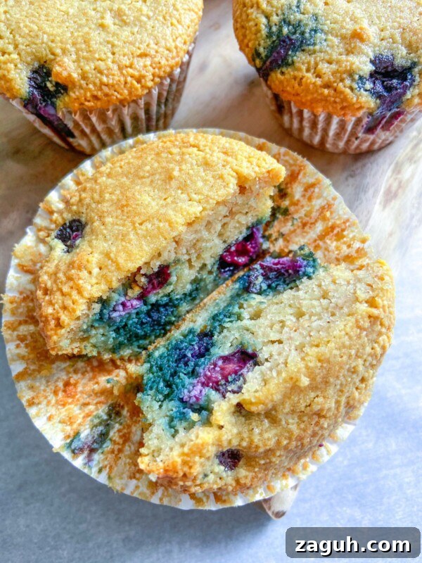Moist Pumpkin Spice Muffins with Simple Ingredients 14 A close-up of Almond Flour Blueberry Muffins cut in half, revealing a moist interior with juicy blueberries. Whole muffins in paper liners rest on a wooden surface in the background.