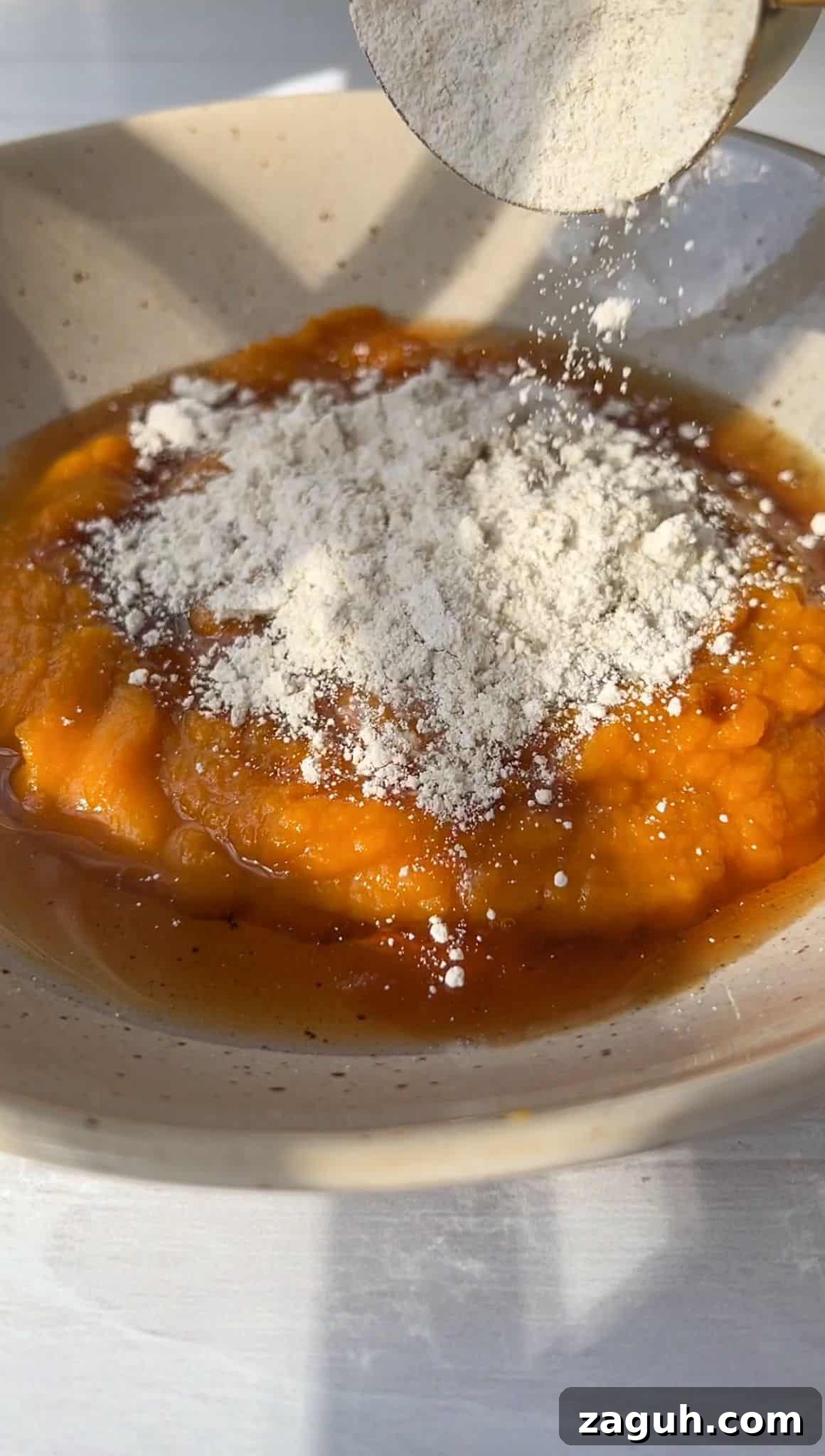 Moist Pumpkin Spice Muffins with Simple Ingredients 4 A bowl contains orange pumpkin puree topped with a spoonful of white flour. Warm sunlight casts soft shadows, highlighting the rustic texture of the brown-speckled ceramic bowl and the smooth surface of the puree.