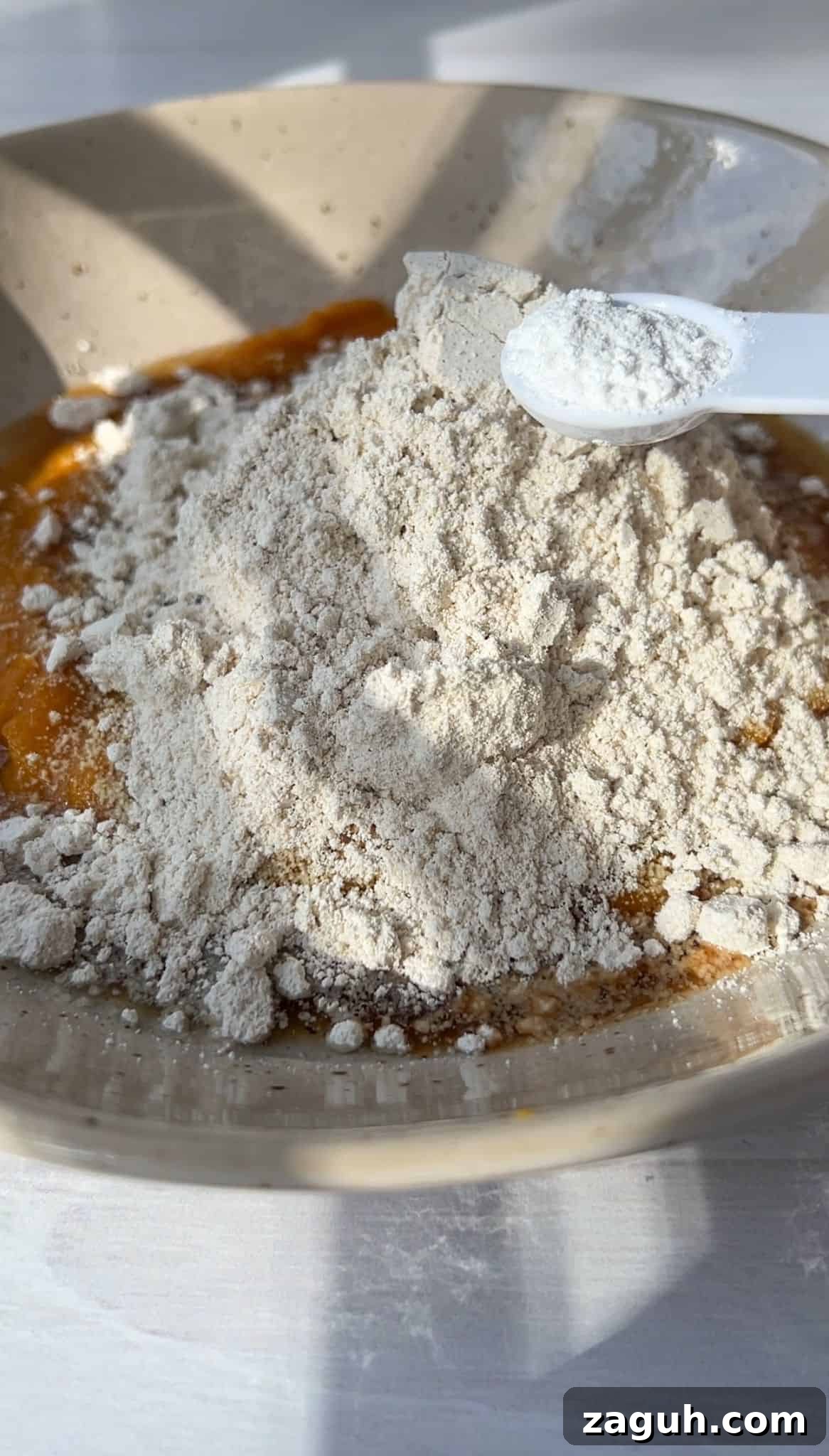 Moist Pumpkin Spice Muffins with Simple Ingredients 5 A bowl filled with a mixture of flour on top of a liquid, possibly dough or batter, with a measuring spoon resting on the flour. Sunlight casts shadows, highlighting the powdery texture of the flour.