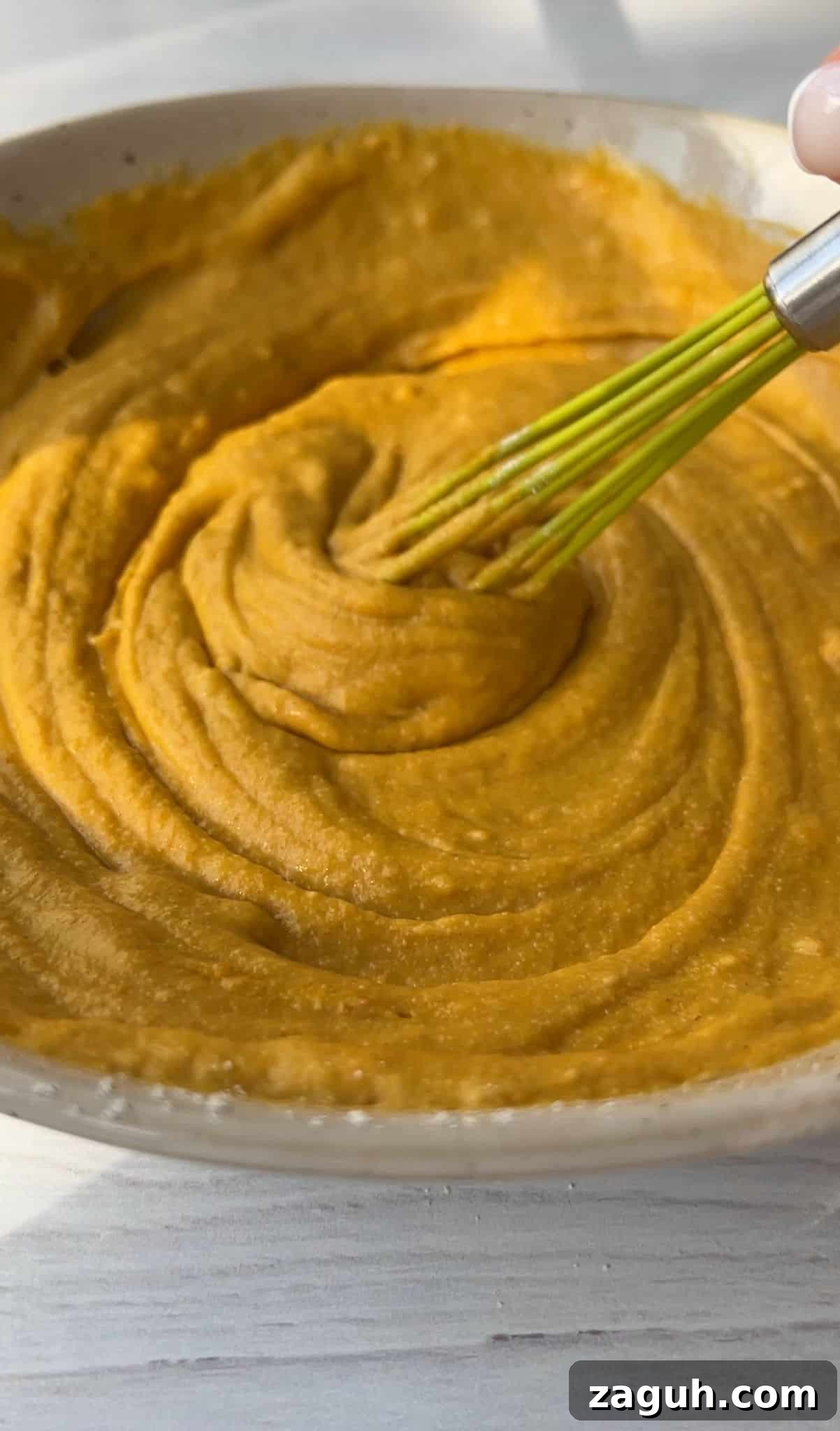 Moist Pumpkin Spice Muffins with Simple Ingredients 6 A close-up of a creamy, orange-hued mixture being whisked in a beige bowl. The texture appears smooth and thick. The bowl is placed on a light wooden surface.