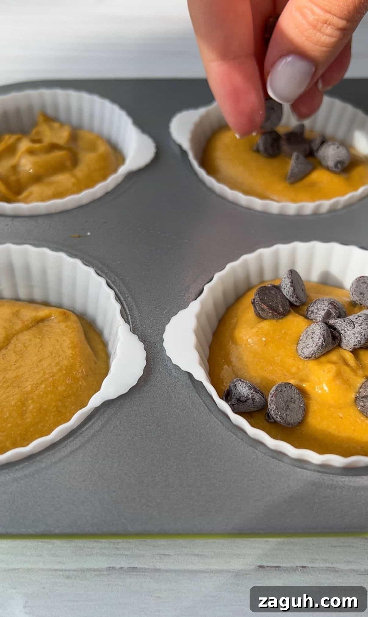 Moist Pumpkin Spice Muffins with Simple Ingredients 7 A hand sprinkles chocolate chips over batter in silicone muffin cups placed on a baking tray. The batter is a golden color, filling four cups, ready for baking.