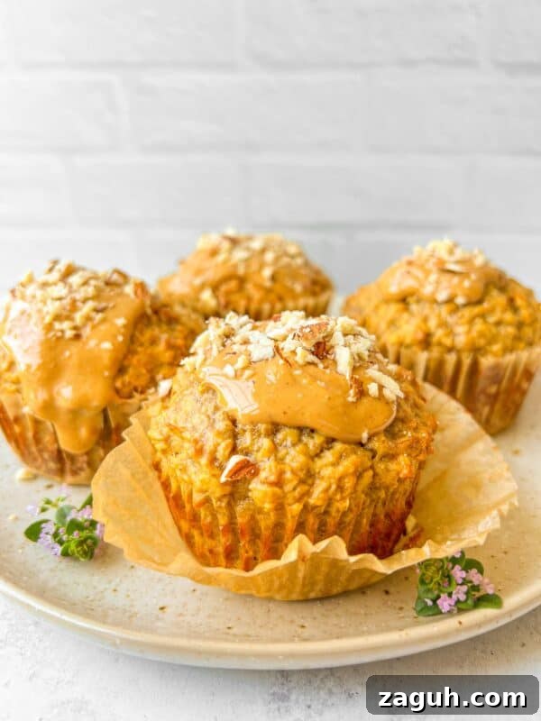 Moist Pumpkin Spice Muffins with Simple Ingredients 10 Four muffins on a plate, each topped with a glossy spread and sprinkled with chopped nuts. The plate is garnished with small purple flowers. The background is a white brick wall, adding a rustic touch.