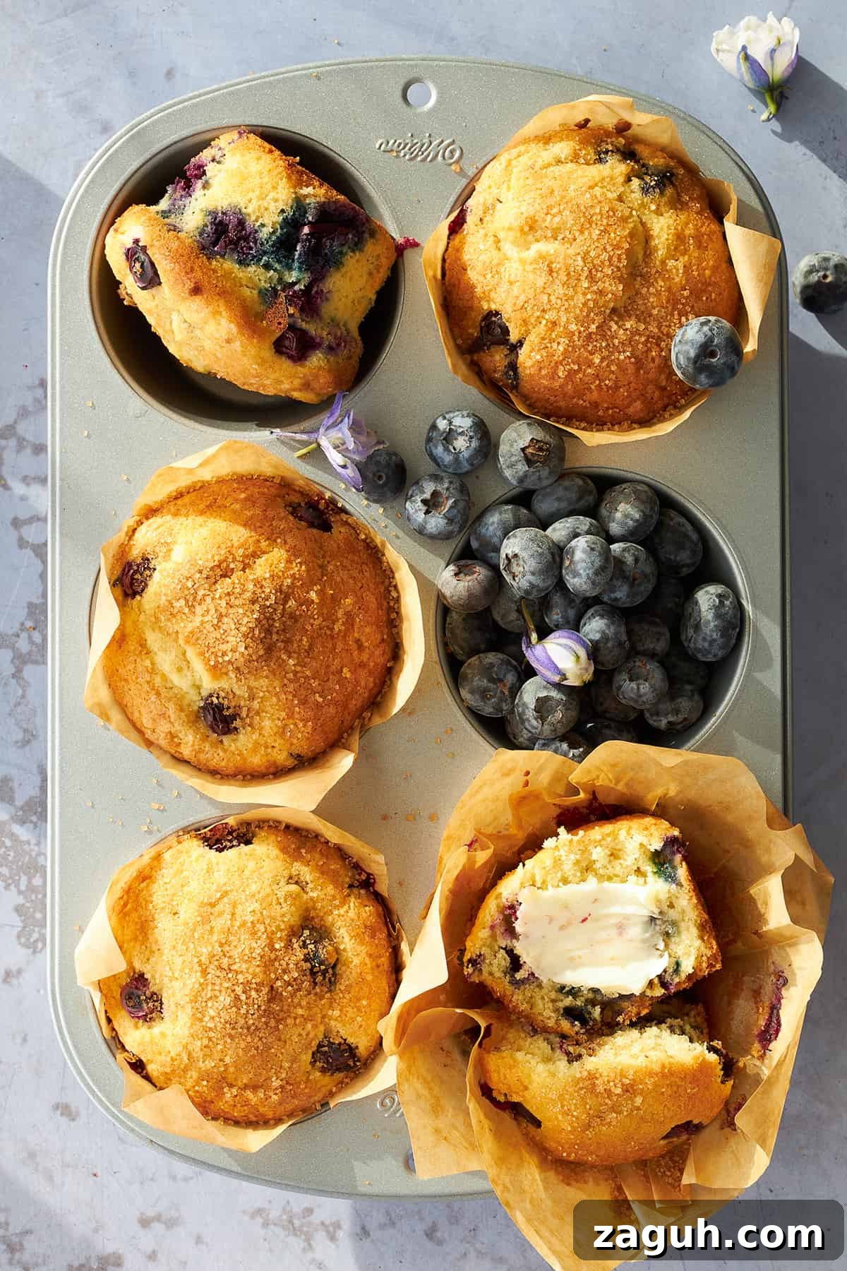 Starbucks-Style Blueberry Muffins Recipe: Make Them at Home 3 Baked blueberry muffins in a muffin tin, one sliced and topped with butter.
