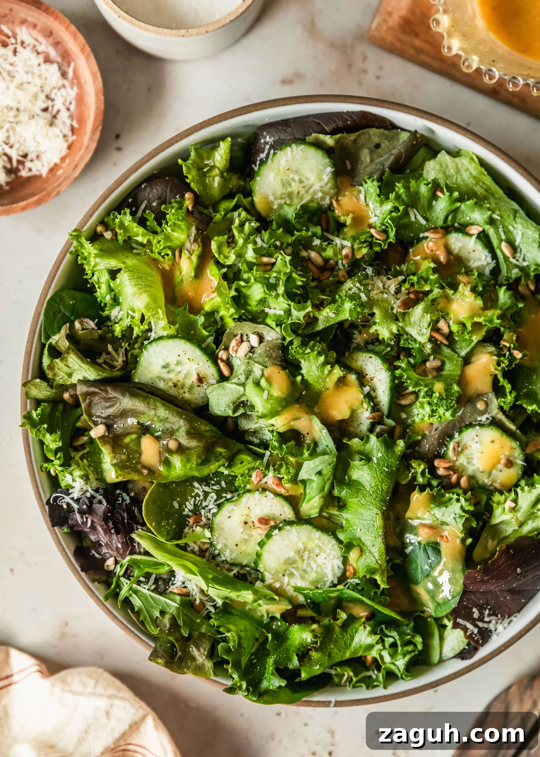 Green Salad with Sherry-Shallot Vinaigrette Recipe 2 A stoneware bowl of green salad with sherry shallot dressing on a tan counter next to a white linen, glass jar of dressing, and wood bowl of parmesan.