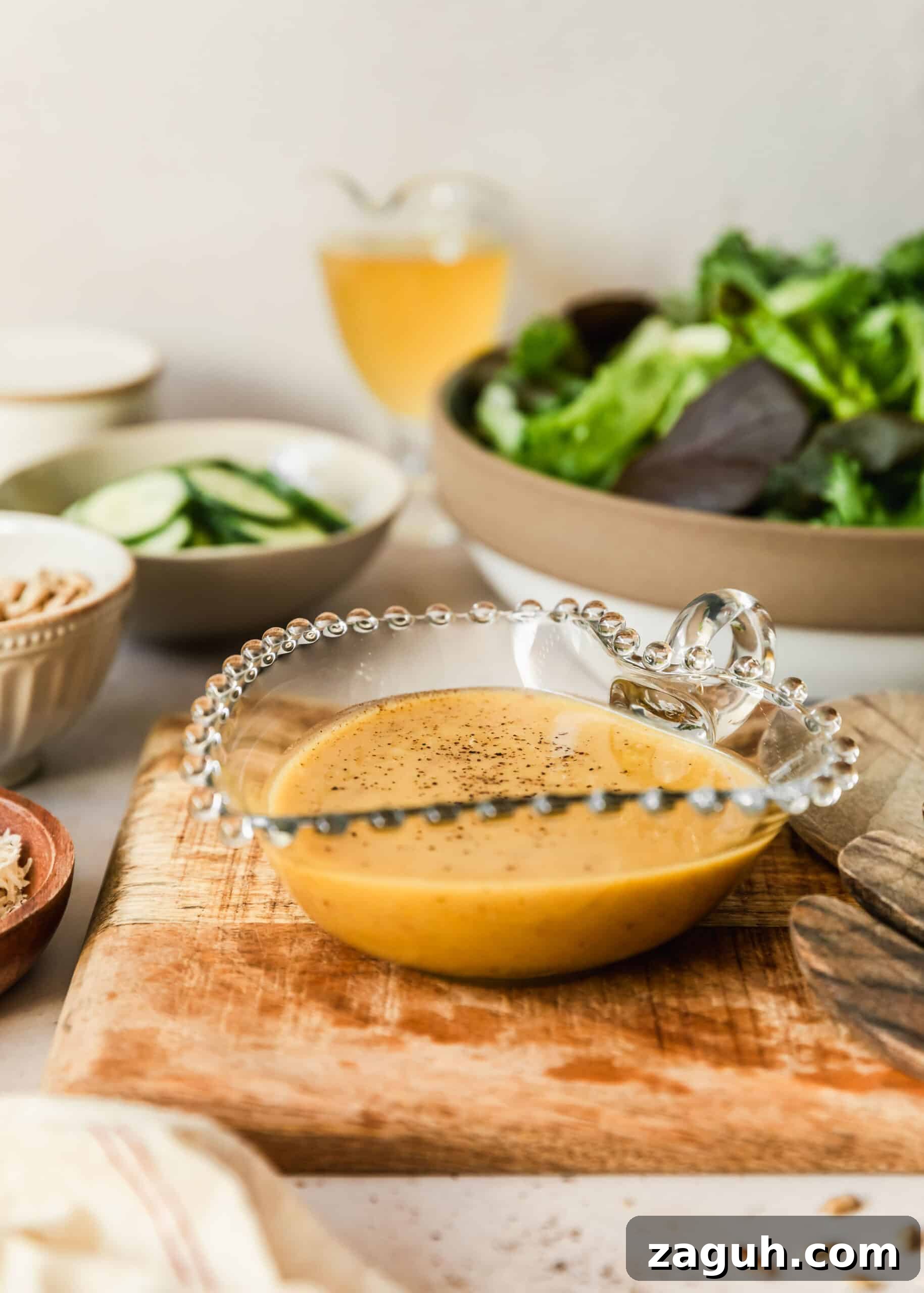 Green Salad with Sherry-Shallot Vinaigrette Recipe 7 A glass container of sherry shallot dressing on a wood board next to white bowls of lettuce, cucumbers, and sunflower seeds on a tan counter.