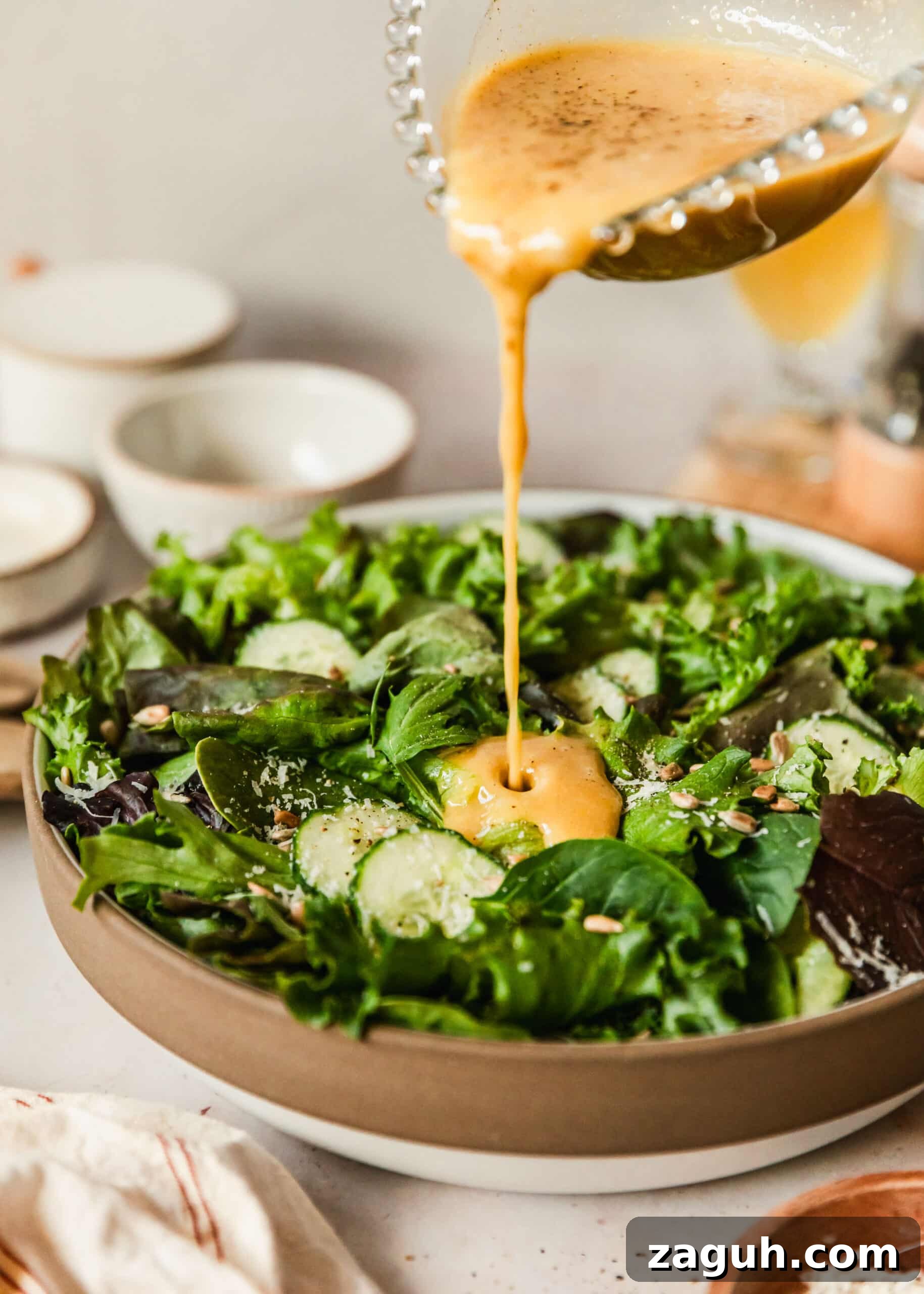 Green Salad with Sherry-Shallot Vinaigrette Recipe 8 A hand pouring a glass container of sherry shallot vinaigrette into a stoneware bowl of lettuce on a tan counter next to white bowls and a white linen.