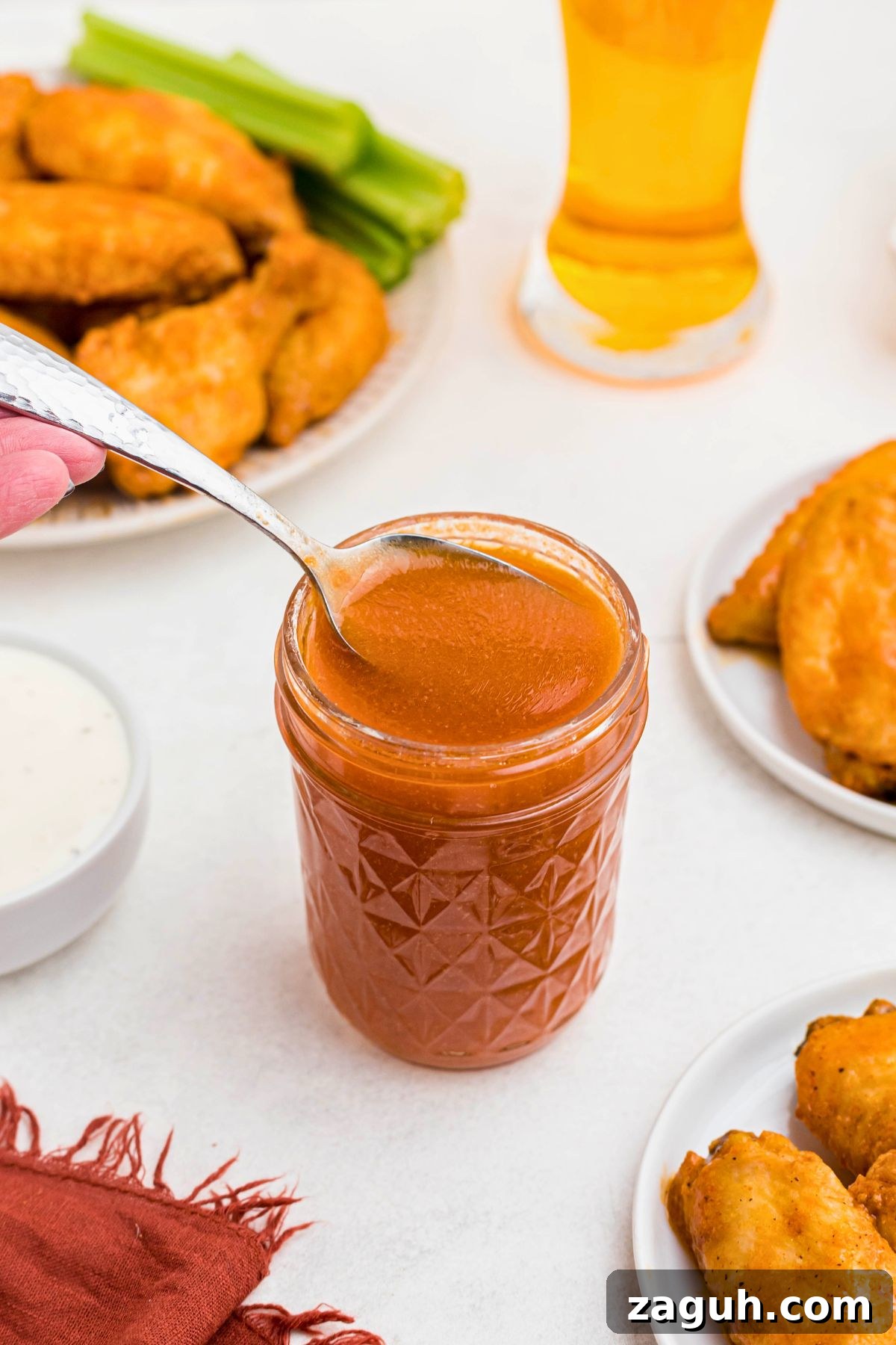 Classic Buffalo Sauce Recipe: Spicy Wing & Dip Sauce in Minutes 7 Orange sauce in a small jar, being spooned out, in front of plates of chicken wings.
