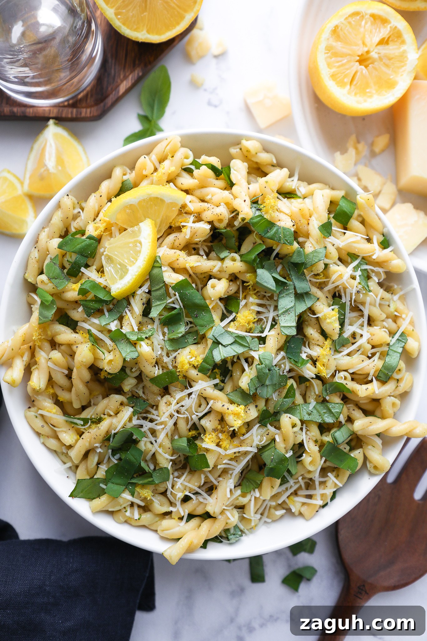 Lemon Basil Pasta Salad with Creamy Dressing and Fresh Herbs 3 Lemon basil pasta salad in a white bowl surrounded by wooden serving utensils, parmesan cheese and lemon slices