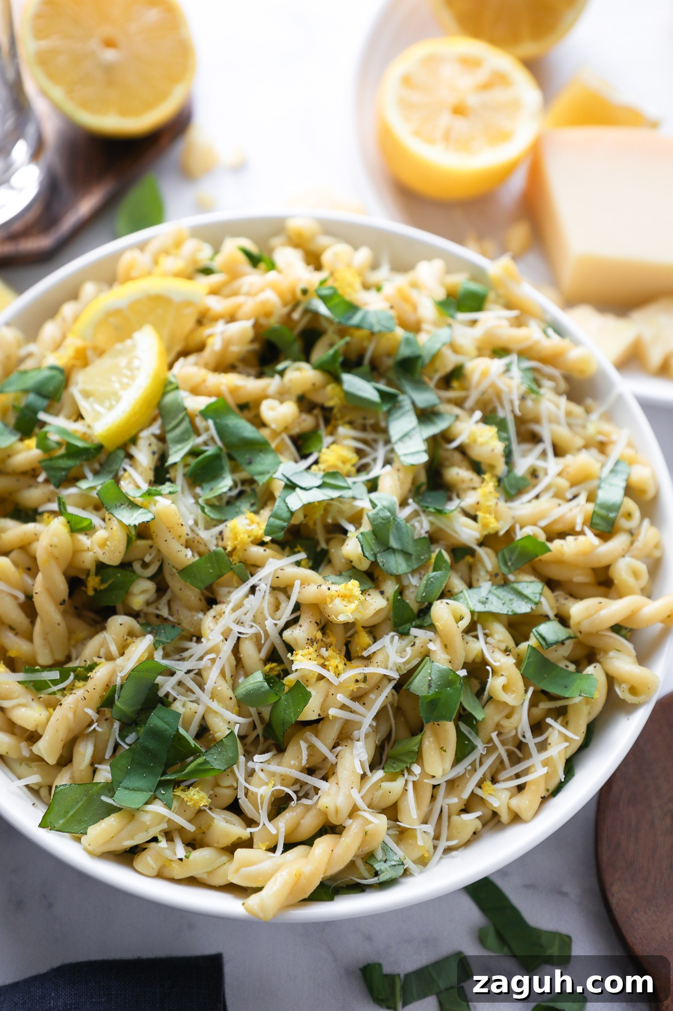 Lemon Basil Pasta Salad with Creamy Dressing and Fresh Herbs 4 Angle view of a bowl of lemon pasta salad sprinkled with fresh lemon zest, basil and shredded parmesan cheese sitting next to lemons cut in half