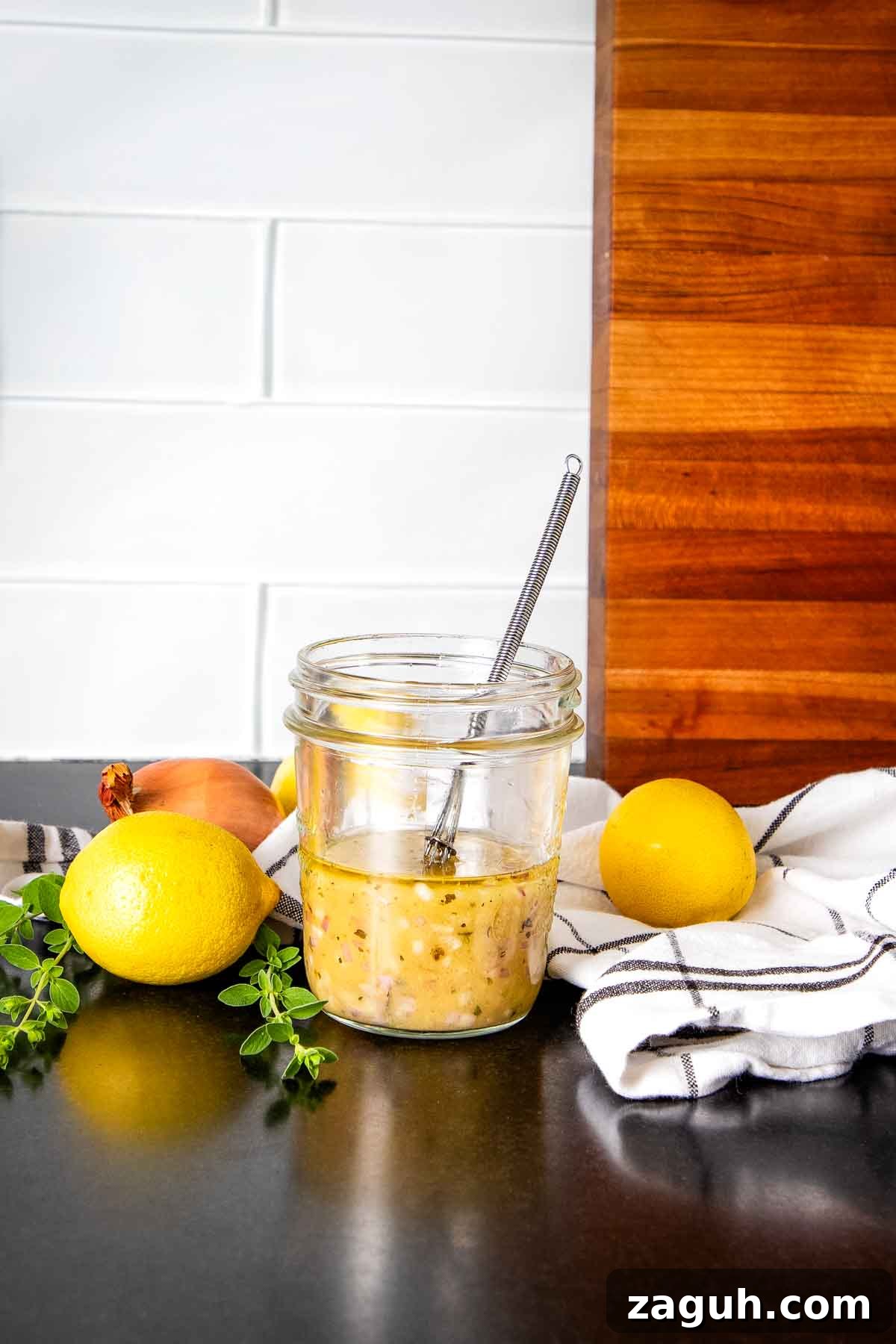 Yellowish salad dressing in a jar with a whisk and some lemons and fresh herbs surrounding on the counter.