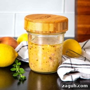 Glass jar with wooden lid filled with salad dressing, surrounded by fresh lemons, a shallot and some fresh herbs.