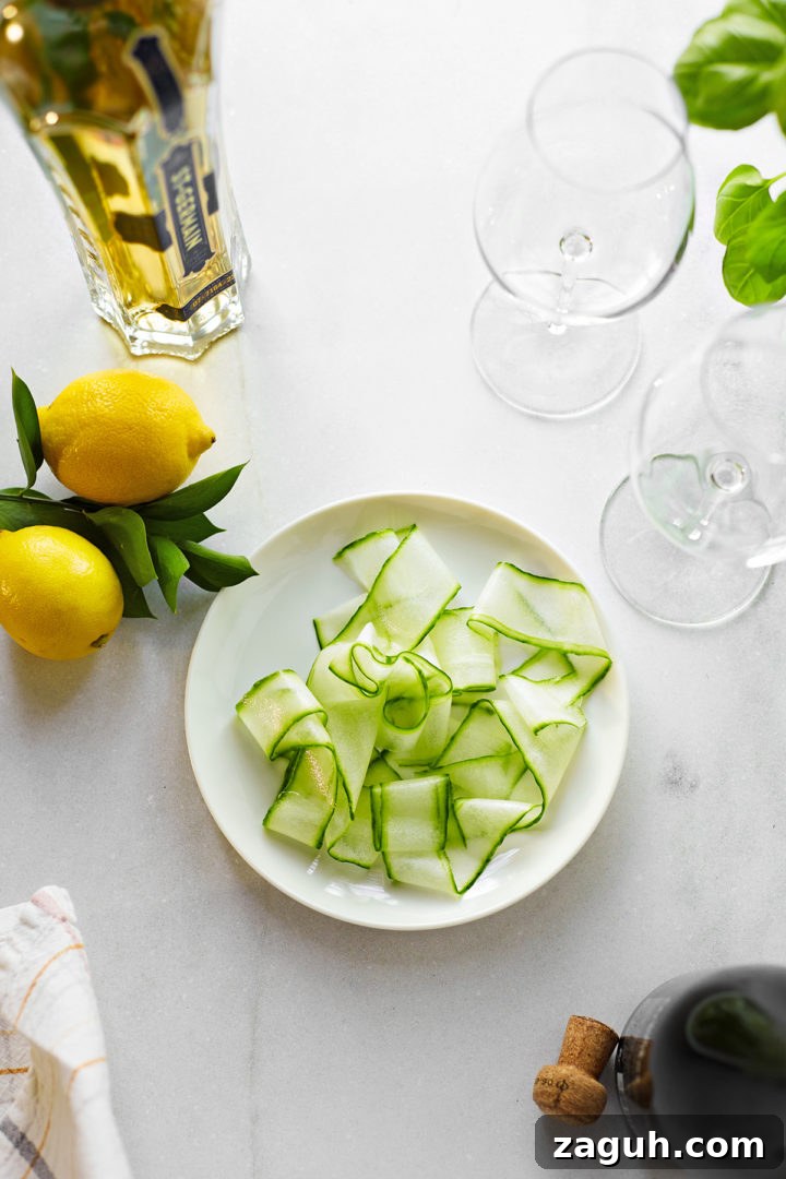 St-Germain Spritz Cocktail Recipe: How to Make a Bright French Spritz 4 sliced cucumber ribbons and fresh lemons next to a bottle of st germain and wine glasses on a marble counter