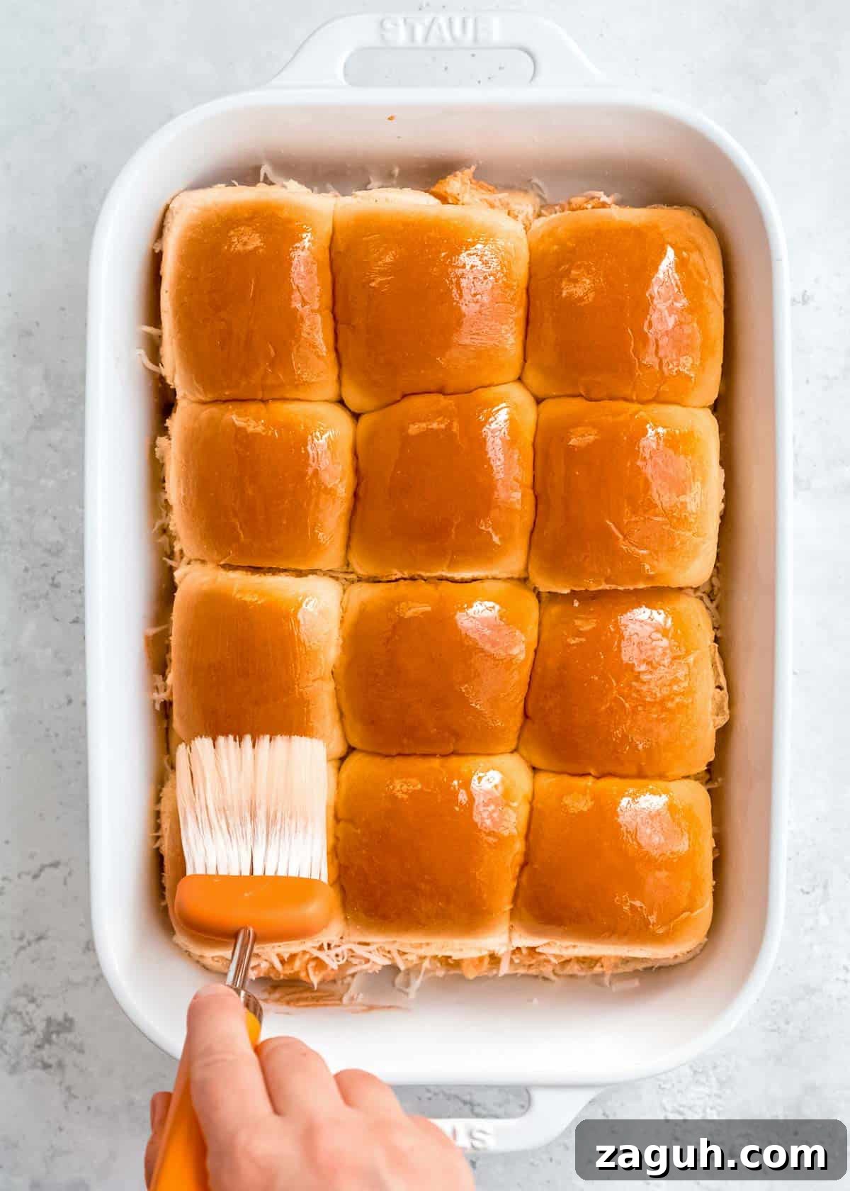 Spicy Buffalo Chicken Sliders with Blue Cheese Slaw 11 top buns added to the white casserole dish; a hand using a brush to coat the top of the buns with melted butter