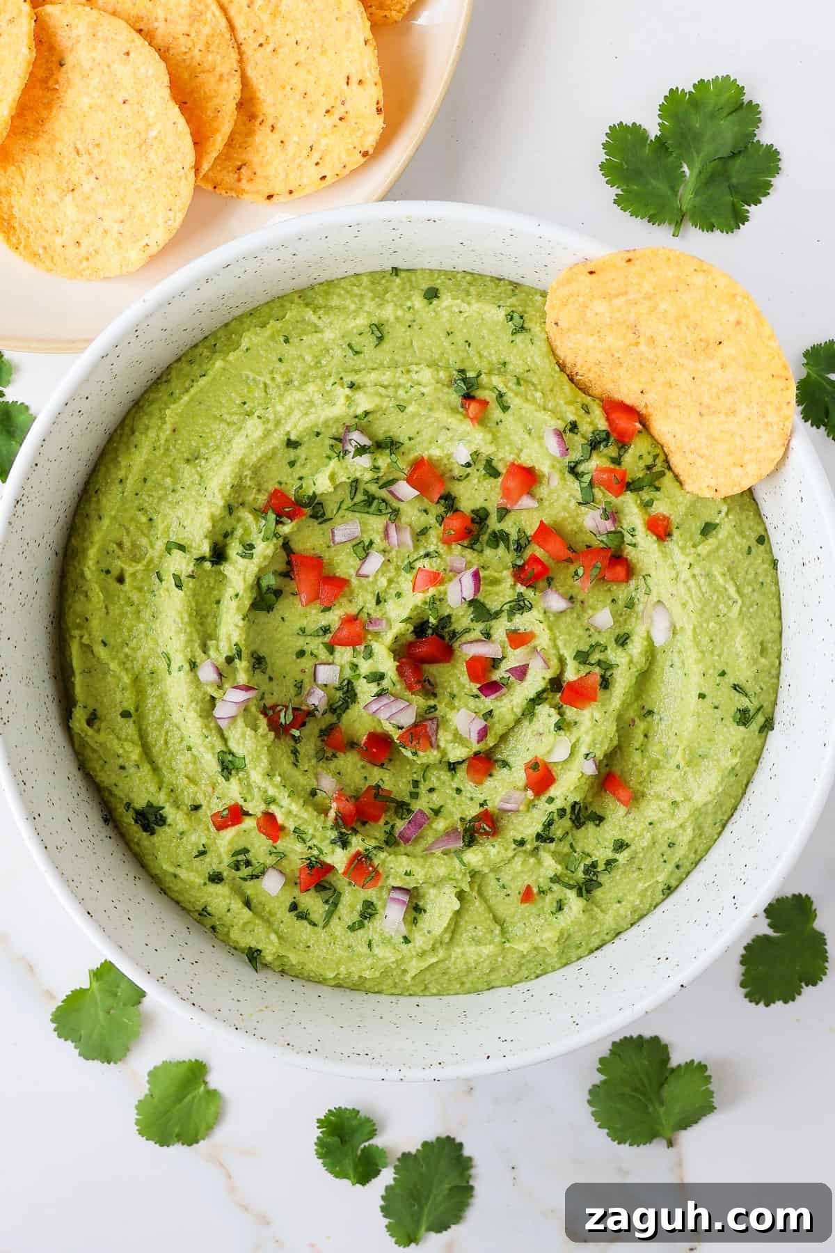Protein-Packed Edamame Guacamole Recipe for Muscle Fuel 2 Guacamole in a bowl with a tortilla chip plus diced tomatoes and onions on top.