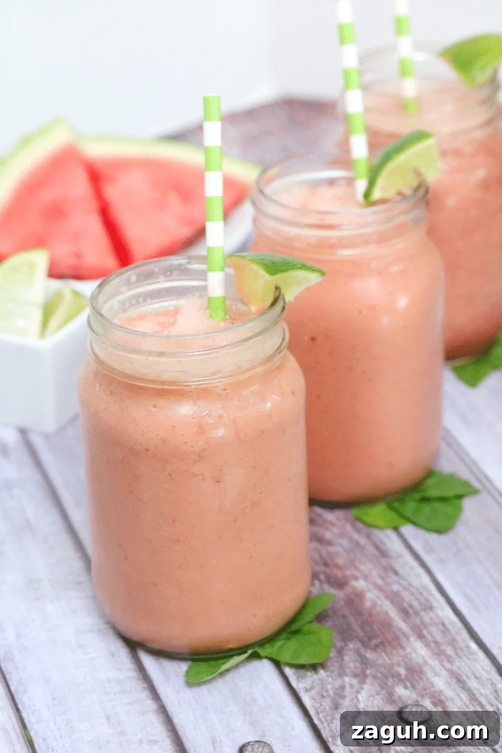 Dairy-Free Watermelon Protein Smoothie Recipe for Summer Energy 2 Three mason jars filled with watermelon smoothie and topped with green straws and lime sitting on mint leaves and fresh watermelon behind them