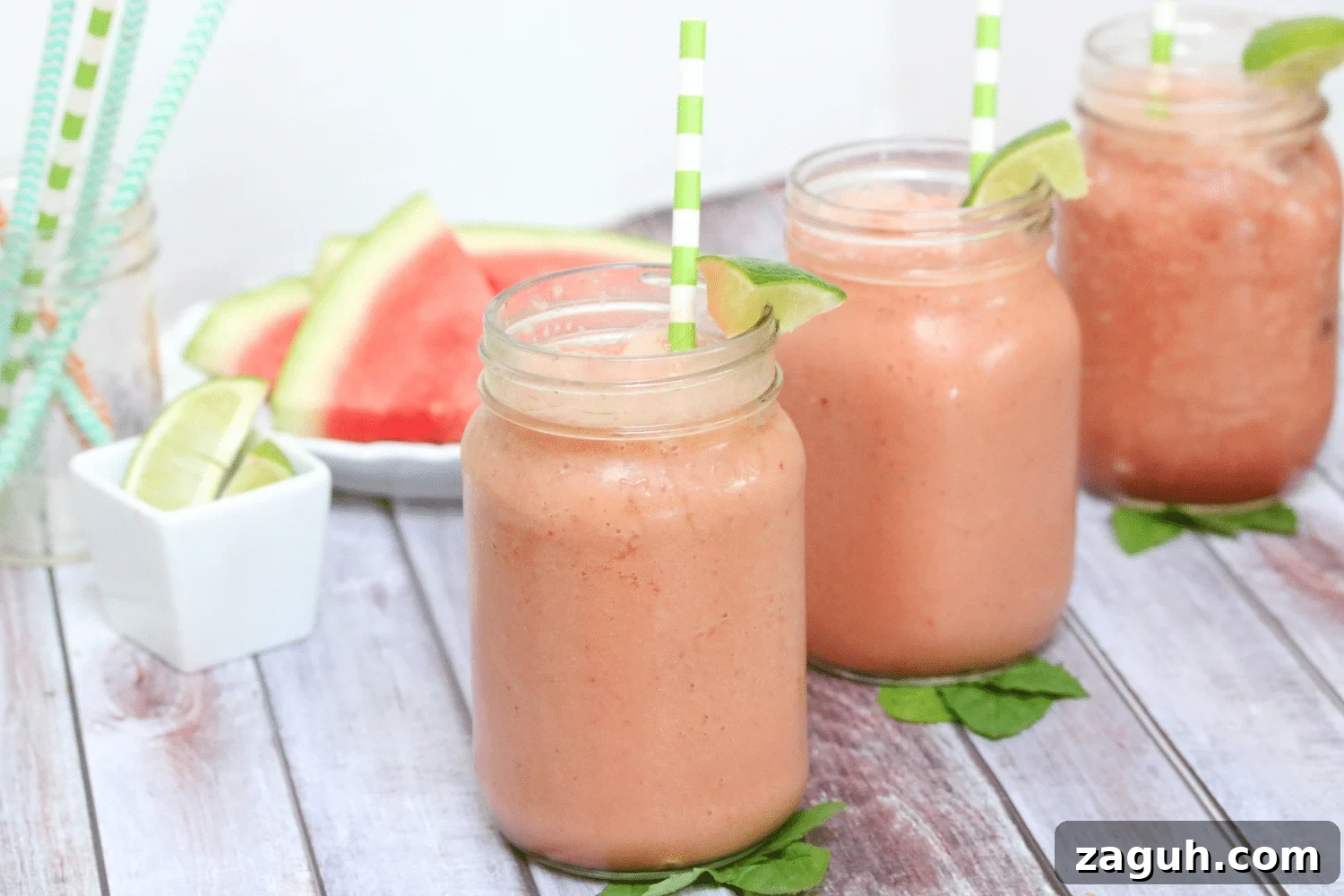 Dairy-Free Watermelon Protein Smoothie Recipe for Summer Energy 3 Three mason jars lined up filled with a pink watermelon mint smoothie topped with cut lime and a green straw in each and sliced watermelon behind them